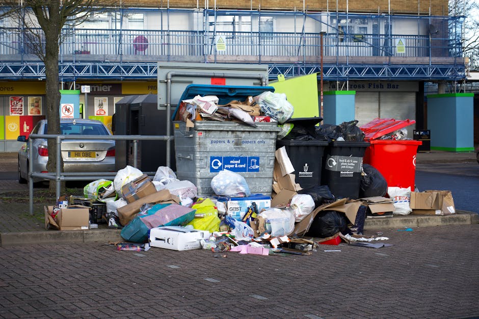 A large collection of mixed household waste and rubbish is piled on a paved urban street, including cardboard boxes, plastic bags filled with trash, black and red kerbside bins, and various paper and packaging materials. The scene is located in front of a building with a blue metal scaffold structure and signage indicating it may house shops or businesses, with a row of convenience stores at the background. A small silver car is parked adjacent to the litter pile, partially blocked by a metal railing that separates the waste area from the vehicle. The waste appears to be a combination of general rubbish collected for disposal or private clearance, with some items spilling onto the pavement, suggesting an uncollected or improperly managed rubbish situation. The lighting implies an overcast day, with diffuse natural light illuminating the scene evenly. This scene exemplifies typical urban rubbish accumulation that might be addressed through independent waste collection services like those offered by House Clearance Brixton, specializing in efficient rubbish removal in local residential and commercial areas.