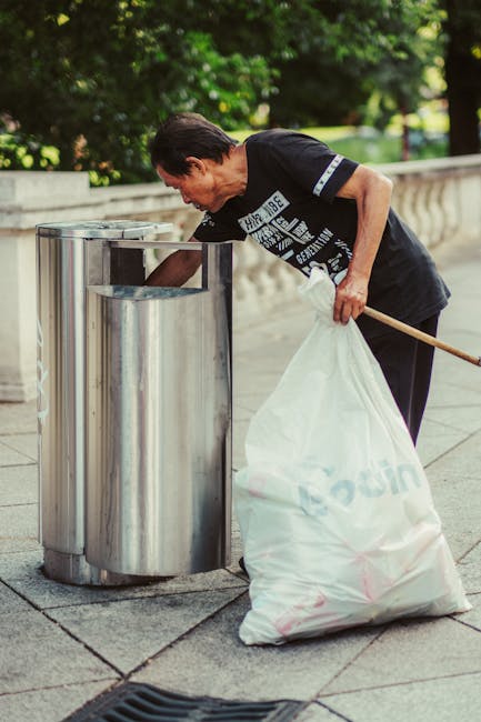 A middle-aged woman with short dark hair, wearing a black t-shirt with white text and dark pants, is seen disposing of rubbish into a stainless steel litter bin on a paved outdoor walkway. She is holding a large white plastic bag filled with waste in her left hand, which rests on her hip, while her right hand is reaching into the bin. She appears to be engaged in a cleaning or rubbish collection activity, possibly part of a community clean-up or regular waste disposal. Surrounding the scene, there are lush green trees and foliage in the background, providing a natural environment that contrasts with the modern metallic bin. The area is well-lit, indicating daytime, and the paved surface shows typical outdoor walkways maintained for pedestrian use. The scene reflects an outdoor rubbish removal activity, aligning with private waste handling or external rubbish clearance services such as those offered by House Clearance Brixton, which specializes in rubbish removal and on-site clearance solutions.
