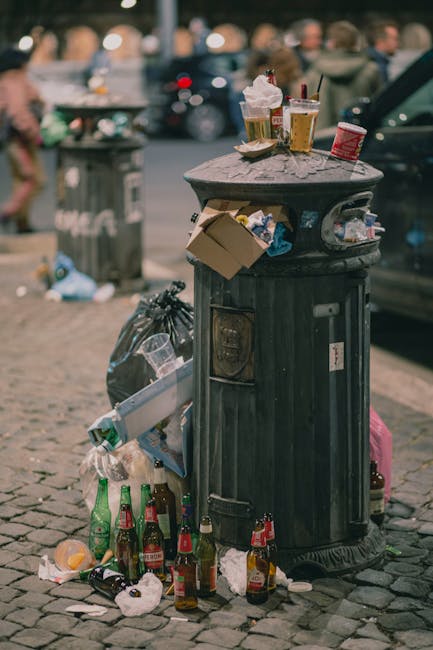 A black, classical-style public rubbish bin made of metal with a domed top, situated on a cobblestone pavement in an urban setting. The bin's surface shows signs of weathering and scratches, and it has a small front access door for waste disposal. The top of the bin is heavily littered with various discarded items, including two half-empty paper cups with plastic lids containing drinks, a clear plastic cup with a straw, and a crumpled paper napkin. In addition, there is a small cardboard box and some crumpled paper on the top of the bin. Surrounding the base of the bin are multiple empty glass bottles, mostly green and brown, some standing upright while others are lying on their sides, along with crumpled paper and plastic waste scattered on the ground. A large black plastic trash bag is leaning against the side of the bin, filled with additional waste. In the background, blurred figures of pedestrians and parked vehicles can be seen, indicating a busy street scene, consistent with urban rubbish disposal and waste management practices in the area. House Clearance Brixton offers services related to waste collection and rubbish removal, often involving on-site clearance that may reduce littering like visible in this scene.
