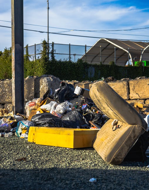 A close-up view of a pile of discarded rubbish on a gravel surface outside, featuring black and yellow trash bags, a large, worn car tire leaning against the pile, and various miscellaneous waste materials. In the background, there is a stone wall and a chain-link fence with green hedges, alongside a tennis court with a white canopy structure and visible fencing, indicating an outdoor urban environment. The scene appears to be an area designated for rubbish collection or private waste disposal, with the presence of waste materials suggesting a service related to rubbish removal. Overhead, power lines cross a partly cloudy blue sky, providing natural lighting that highlights the textures of the waste and surrounding environmental elements. This setup aligns with independent rubbish clearance activities often handled by companies like House Clearance Brixton, highlighting an accessible area for waste collection outside residential or commercial properties.
