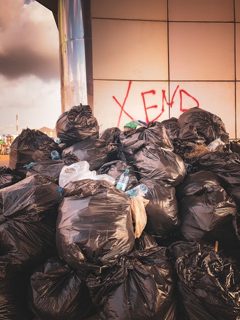 A large pile of black plastic rubbish bags, some tightly tied and others loosely gathered, stacked against a modern building with reflective tiled panels. The bags contain various waste items, with a few plastic bottles visibly protruding from some, indicating general rubbish collection. The surface of the bags appears crumpled and glossy, reflecting ambient light and highlighting their plastic material. Behind the pile, there is red spray-painted graffiti that reads 'XEND' on the building's wall. The surrounding environment suggests an urban setting, possibly on a pavement or alleyway near commercial premises. The scene is lit by natural daylight, with the sky showing dark clouds in the background, contributing a somewhat ominous tone to the overall atmosphere. This image relates to private or independent waste disposal, aligning with services like those provided by House Clearance Brixton, which often handle on-site rubbish removal outside of public waste collection channels.
