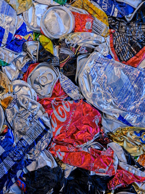 A close-up image displaying a pile of crushed aluminium beverage cans, predominantly silver with some featuring red, blue, black, and gold branding. The cans are deformed from compression, with visible pull-tabs and ridges, scattered and overlapping each other. The surface texture varies between smooth and crinkled, with some cans showing faded or partially visible logos and labels. The background is not visible, suggesting the cans are collected scrap, possibly for recycling or disposal through private waste removal services. The scene emphasizes the importance of rubbish management and the potential for alternative waste handling methods such as on-site clearance or independent collection carried out by companies like House Clearance Brixton, supporting efficient waste disposal in urban areas.