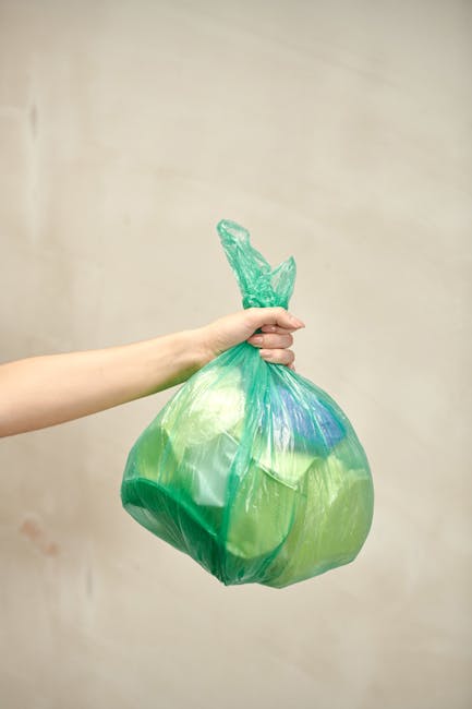 A person's hand holding a green and blue tied plastic rubbish bag, which appears to contain general waste. The bag is slightly translucent, showing some contents inside, and is held firmly at the top with a twist tie. The background is a plain, neutral-toned wall, and the scene is evenly lit, emphasizing the bag's material and colors. This image relates to waste removal services such as private rubbish collection or waste disposal, highlighting an example of waste ready for collection at a residential property. The focus on the bag's texture and the act of holding it suggests the process of rubbish accumulation awaiting professional rubbish removal, as offered by companies like House Clearance Brixton, servicing areas with alternative waste handling options like same-day or on-site clearance for domestic or commercial waste.